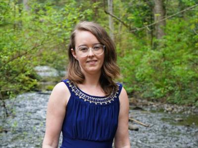 A person in a long dress and glasses standing in front of a river in the forest