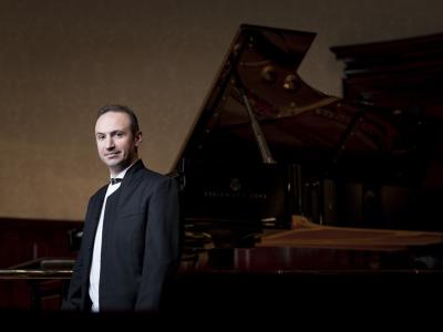 Simon Trpčeski in a formal suit stands in front of a large piano with a straight face and one raised eyebrow