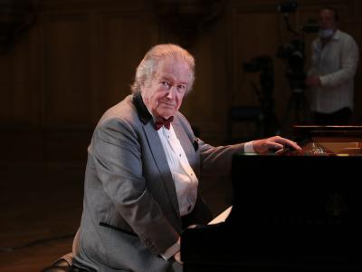 Mikhail Voskresensky in a gray suit, sits at a piano with one hand on top, and turns slightly to look at the camera