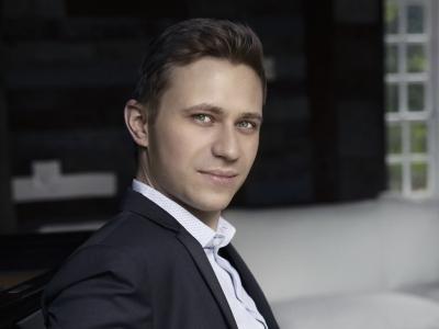 Dmytro Choni in a suit, seated backward at a piano, leaning casually on the keys. Dymtro has an intense gaze and barely noticeable smile.