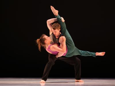 A dancer held horizontally by another. Their eyes are locked and the held dancer's leg is supported straight up in the air by the other dancer