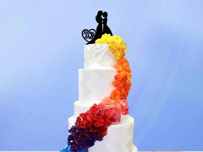 tall tiered white wedding cake with a rainbow cascade of flowers winding down the side, topped with two figures in dresses and a heart that reads Mrs & Mrs.