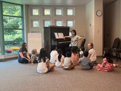 a music instructor in front of a piano surrounded by children sitting on the floor