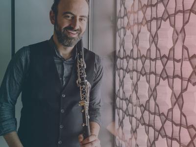 Kinan Azmeh smiling as if mid-chuckle, holding a clarinet next to a stained glass wall