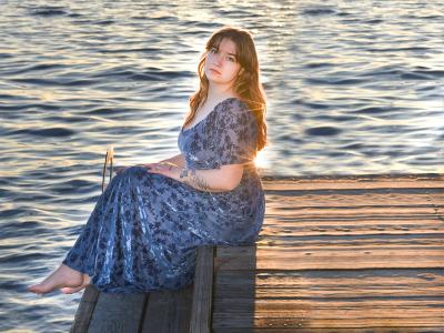 a person in a dress sits on a dock extending into the water with the sun setting in the background