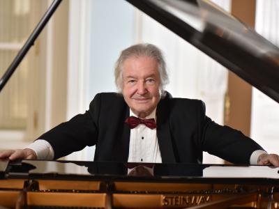 Voskresensky, an elderly man with a friendly face and one lifted eyebrow wearing a nice tuxedo, leans confidently upon a steinway piano