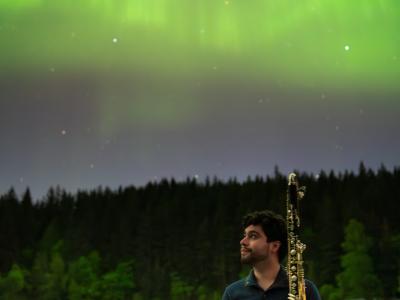 David Bissell standing holding a clarinet, smiling up at a sky filled with aurora borealis
