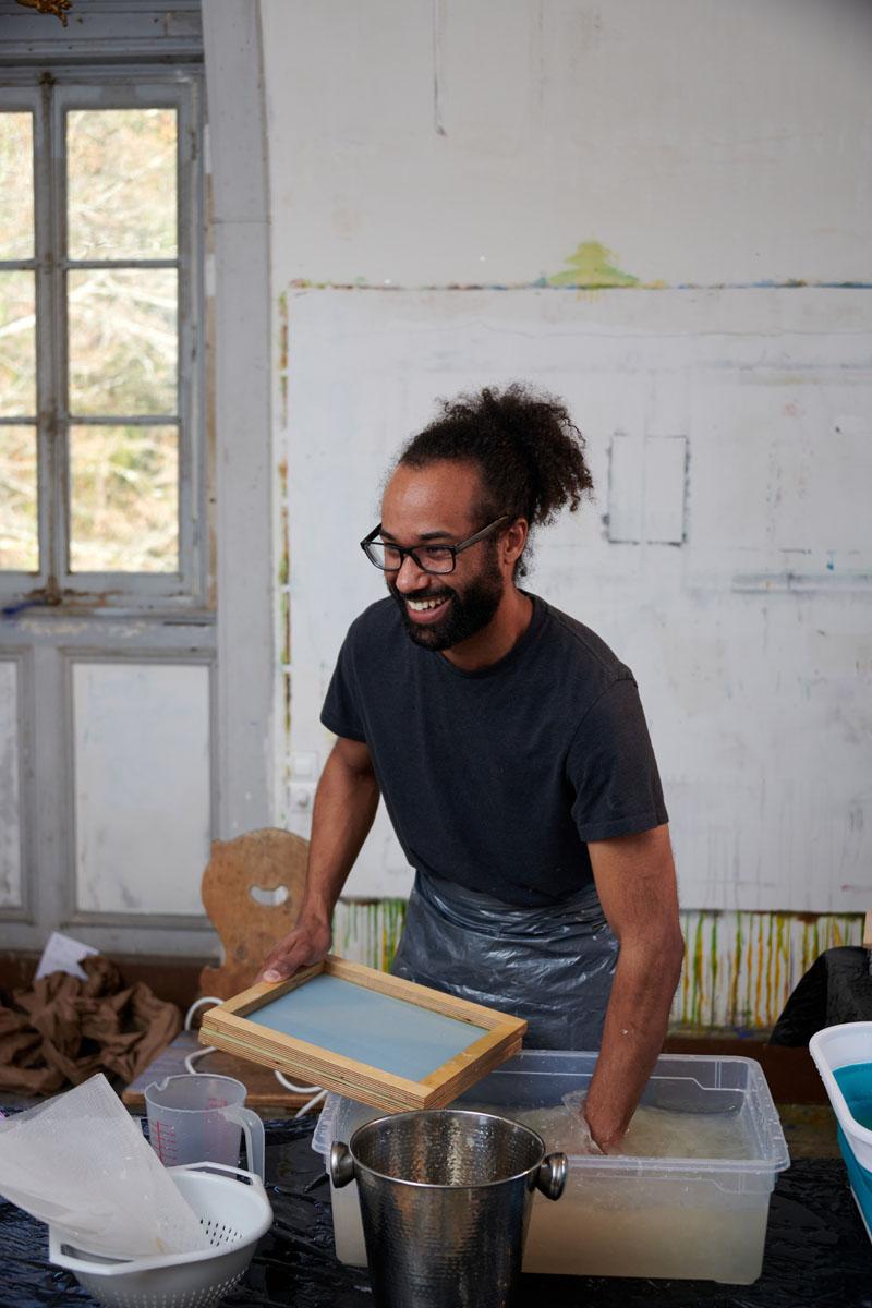 Artist Henry Jackson-Spieker smiles while working in the studio. A paned window in the background.