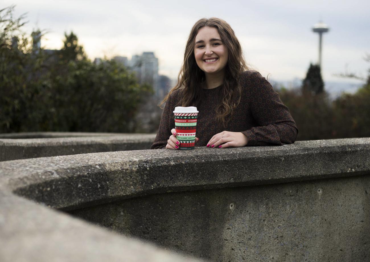 young person leaning on the curb of a fountain smiling holding a decorative paper coffee cup