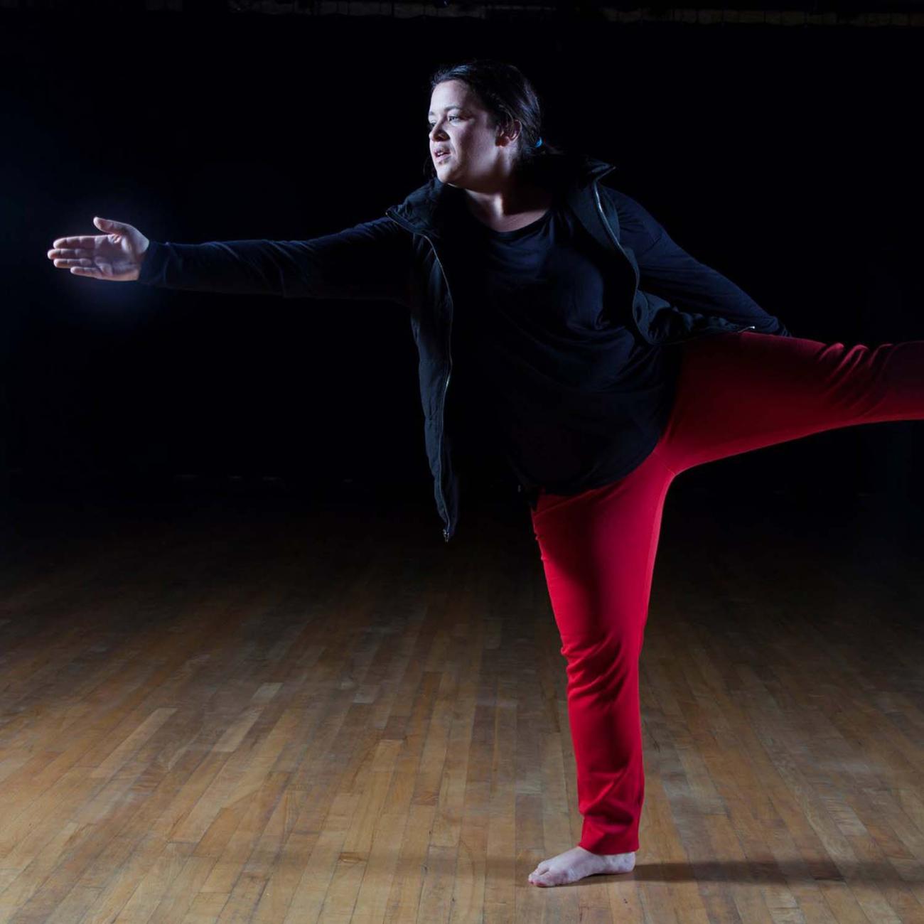a dancer in a dark studio reaches out with extended hand, counterbalanced by a rearward extending leg