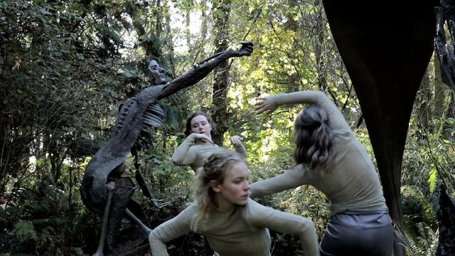 three dancers move in and among large mythical bronze sculptures in a wooded setting