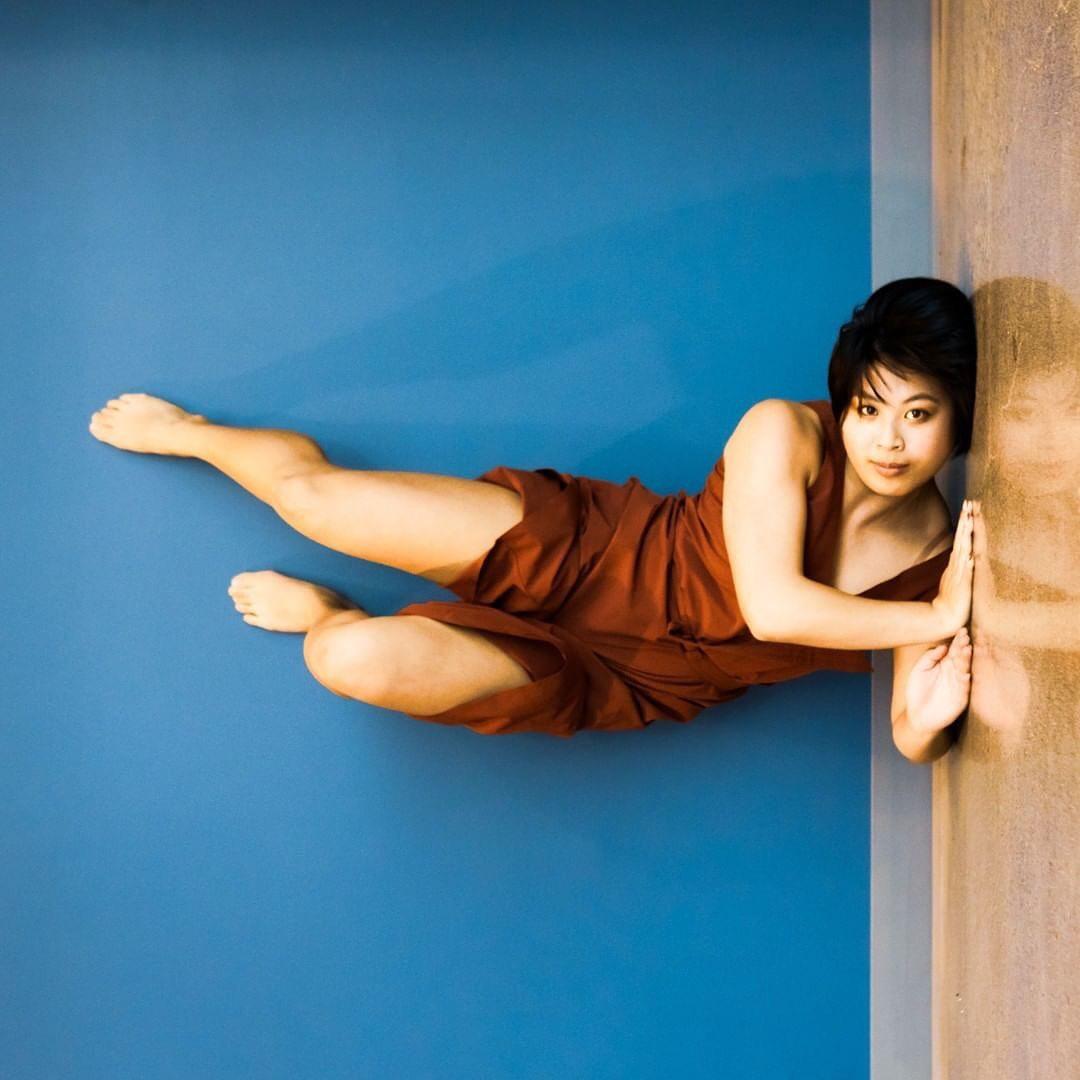 Emma Rose DeSantis appears to be stuck to a wall by her shoulder and neck, but it's a visual trick because the image is rotated 90 degrees on pupose, and she's really in a shoulder stand on the floor. The wall is blue, her short dress is burgundy.
