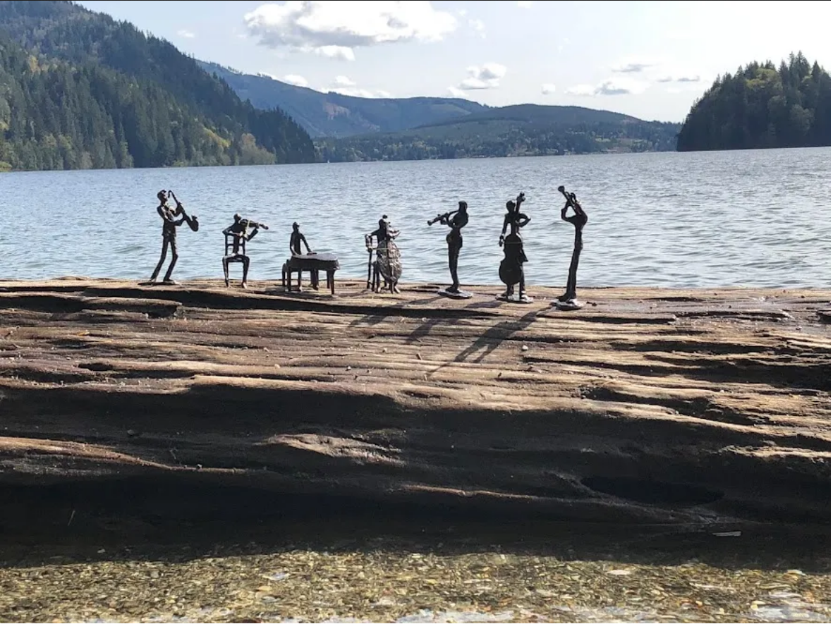 a sextet of small musician figurines sit on a driftwood log near a lake with mountains behind