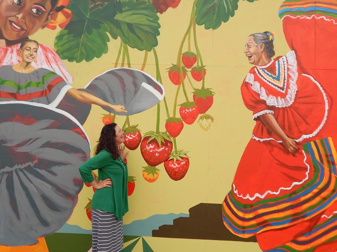 a woman stands in front of a brightly painted mural of Mexican folk dancers