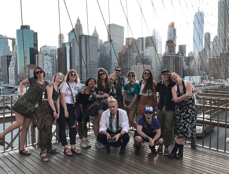 a group of students pose for a photo on their trip to New York City. They are on a bridge with the cables nearby and the skyline behind.