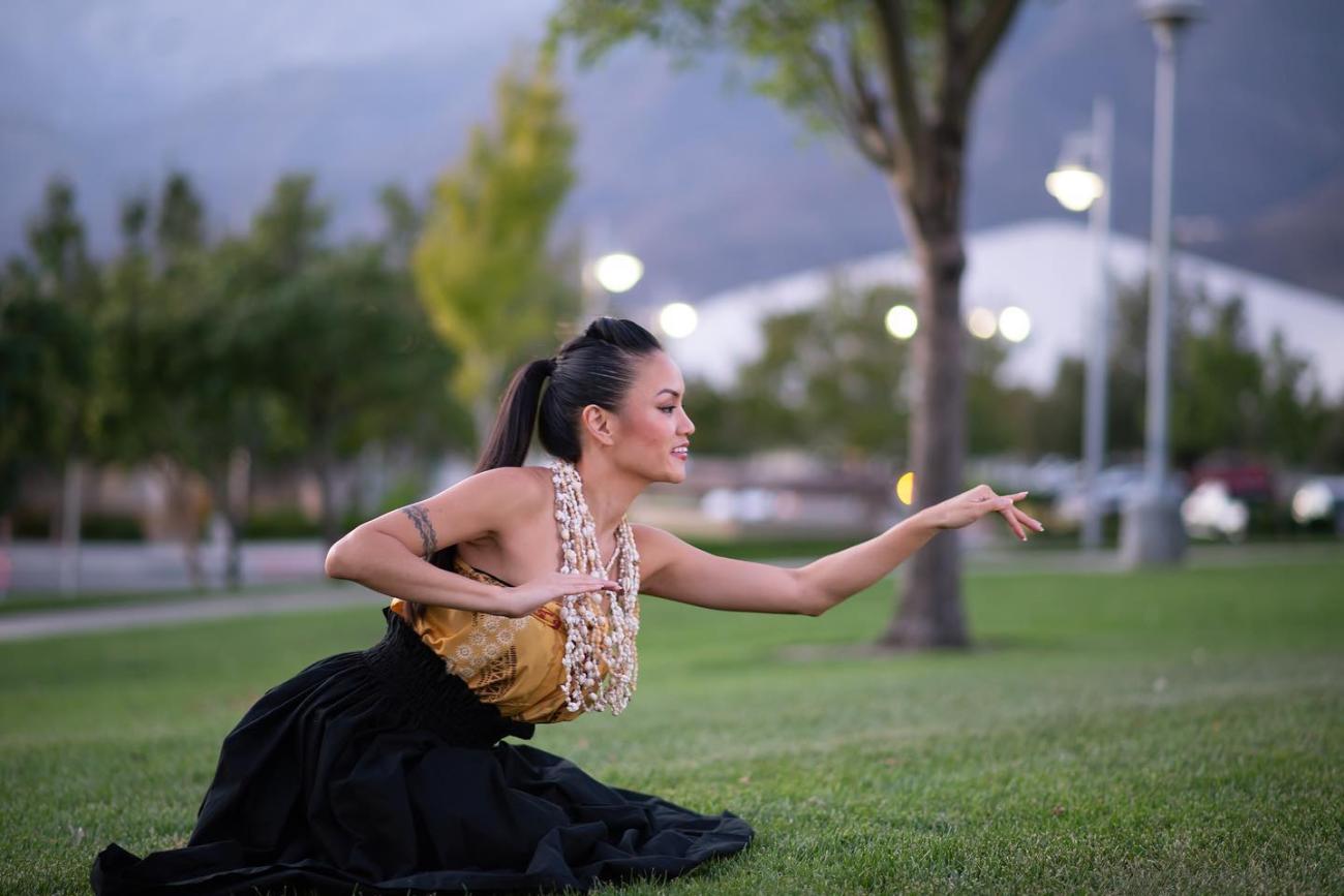 a hula leader kneeling in a dance pose on the grass extending one arm in a graceful gesture