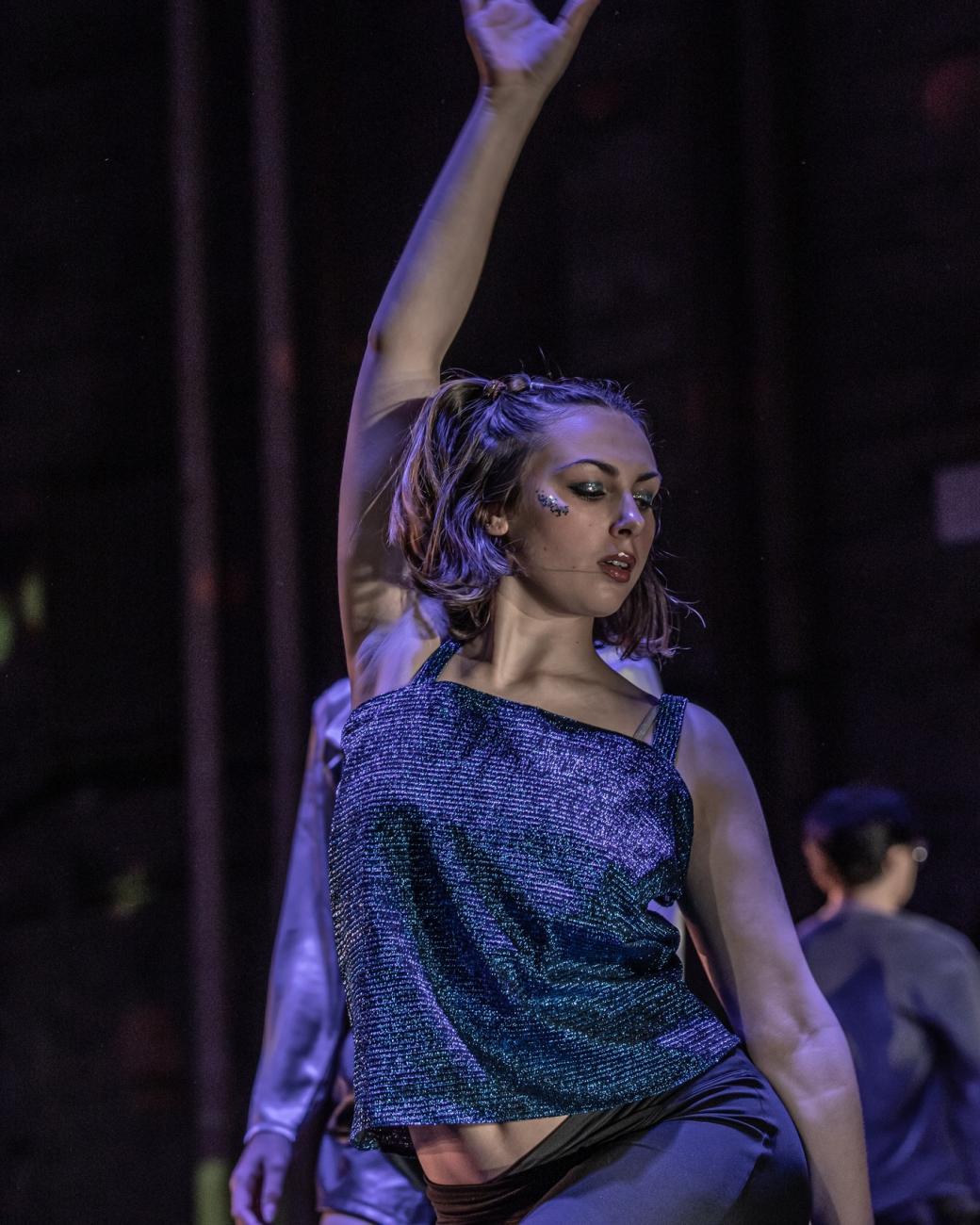 a dancer tossing her right arm in the air like she is grooving at a supa cool techno club, wearing a shiny blue metallic halter top and eyeshadow that just won't STOP glimmering under the lights