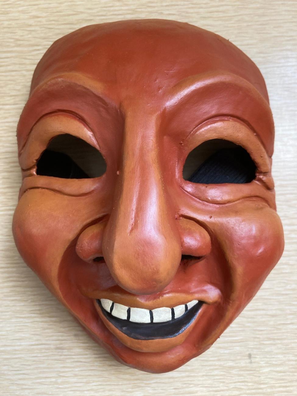 Earth-colored cartoonish laughing mask, with a big nose and empty eye sockets