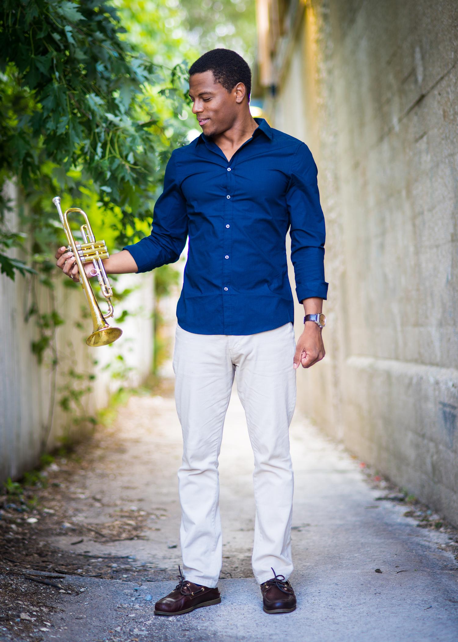 Justin Copeland standing outdoors, smiling at the trumpet in his hand