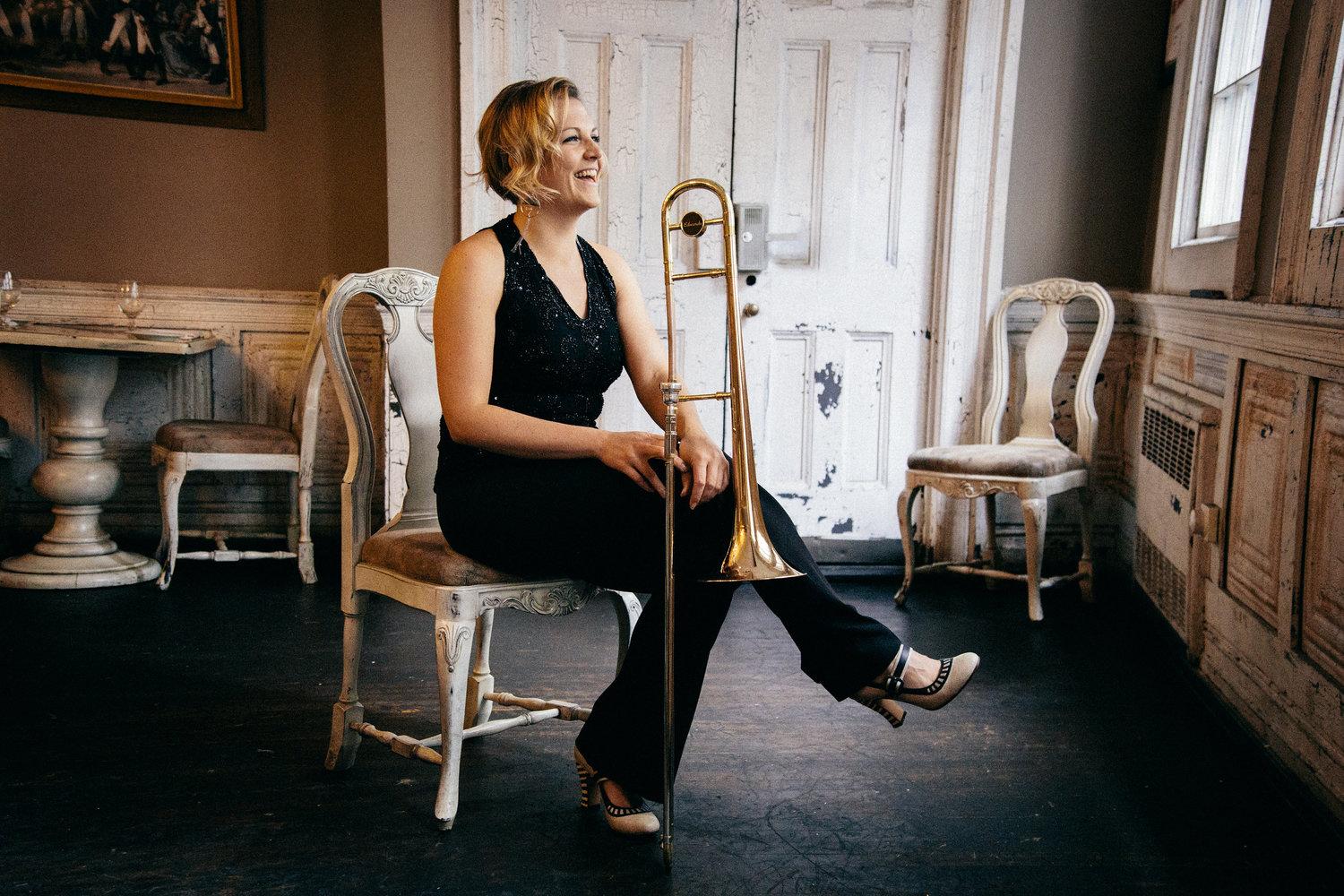 A person wearing a fancy, modern outfit sitting in a chair in a distressed, Victorian style room, holding a trombone and laughing joyfully
