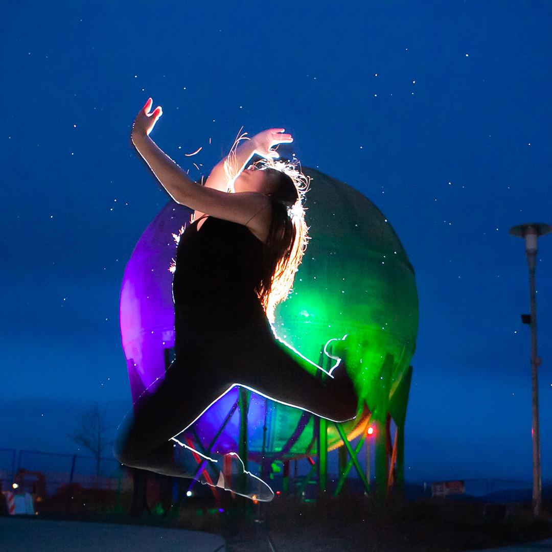 A dancer mid-leap, arched back, arms and legs curled. Behind a colorful sphere structure glows under a starry sky.
