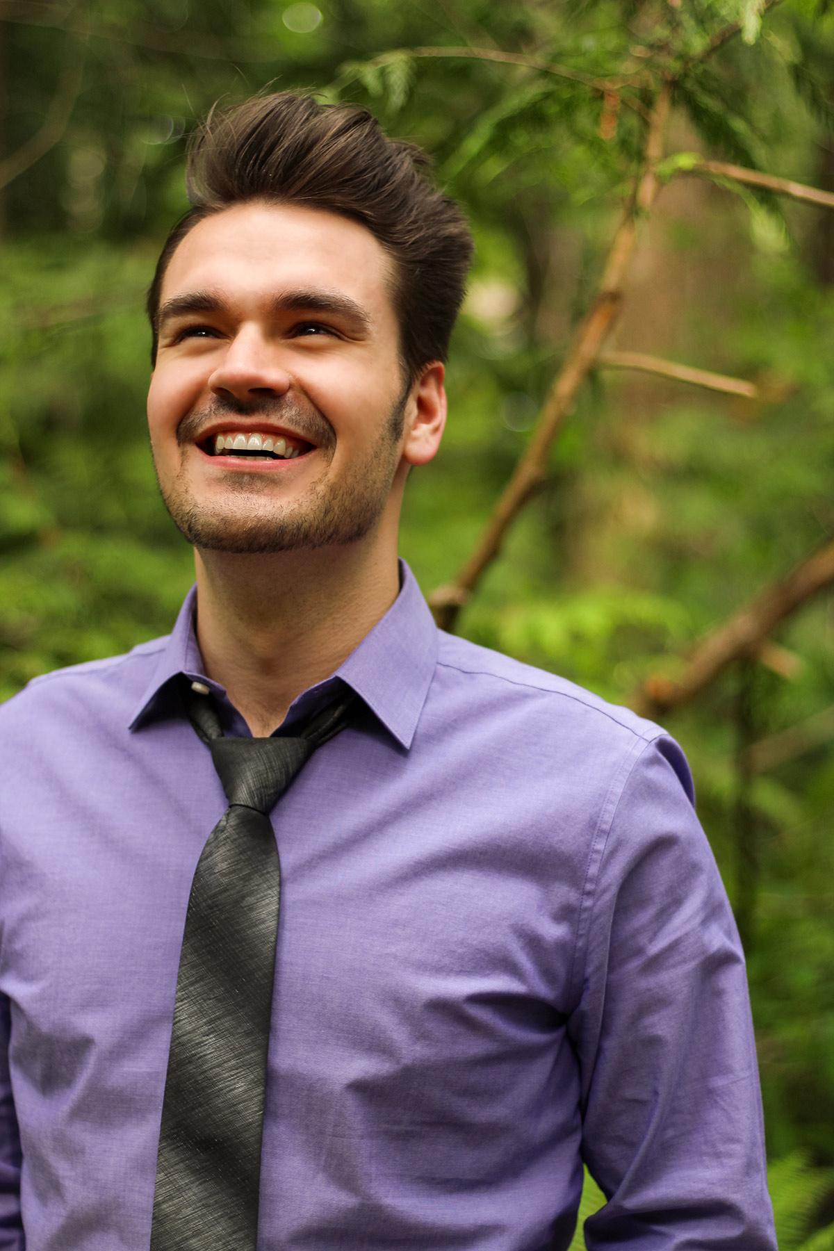 young fellow smiling broadly. Purple collared shirt, top button undone, grey necktie.