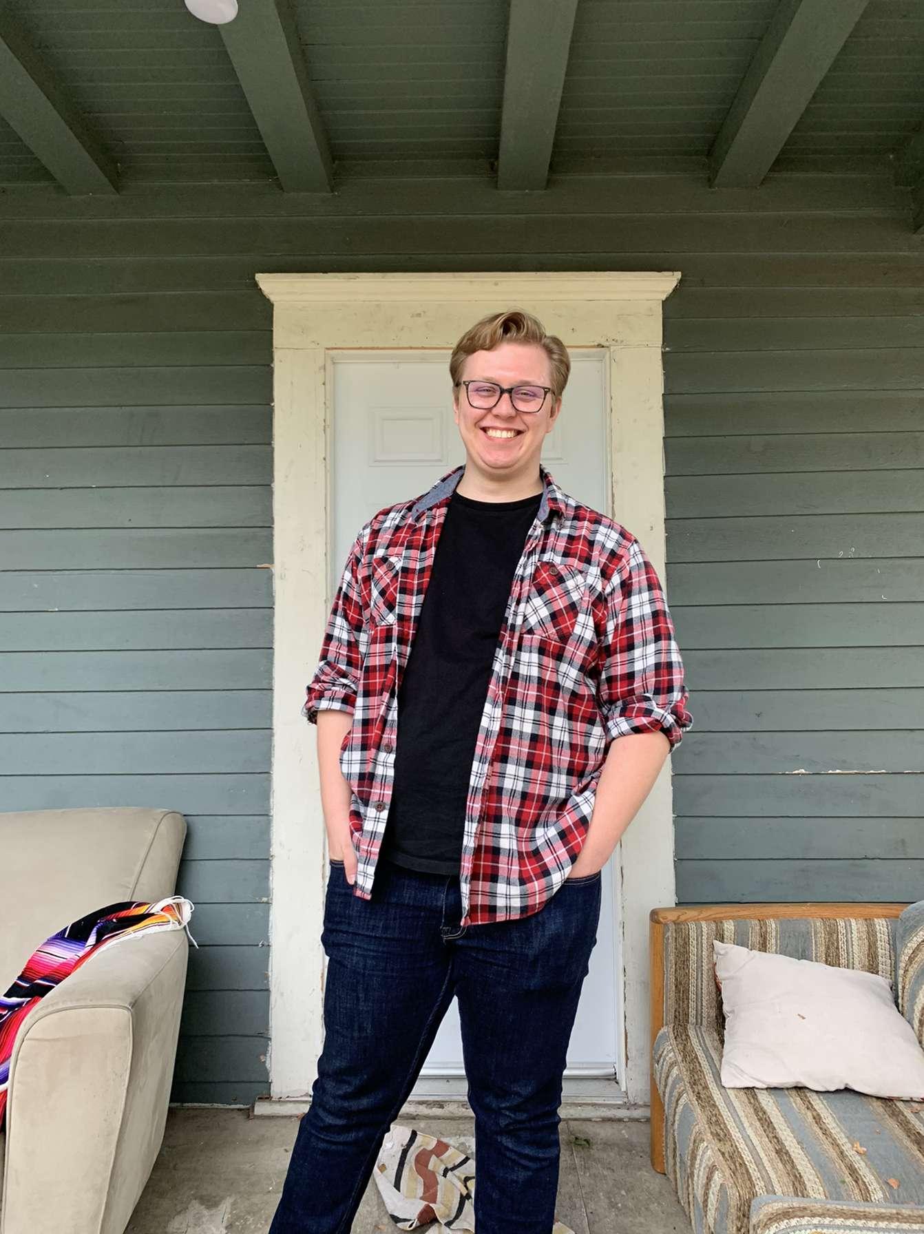 Person in a plaid shirt, unbuttoned, smiling from a gray porch