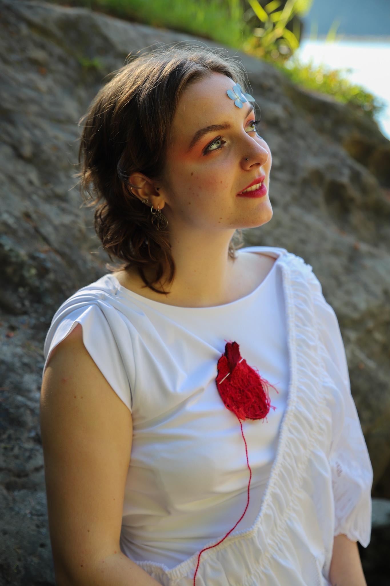 a young performer in a white sleeveless shirt in an outdoor setting. There is something like a anatomical yarn heart attached to the shirt and a butterfly on the person's forehead.