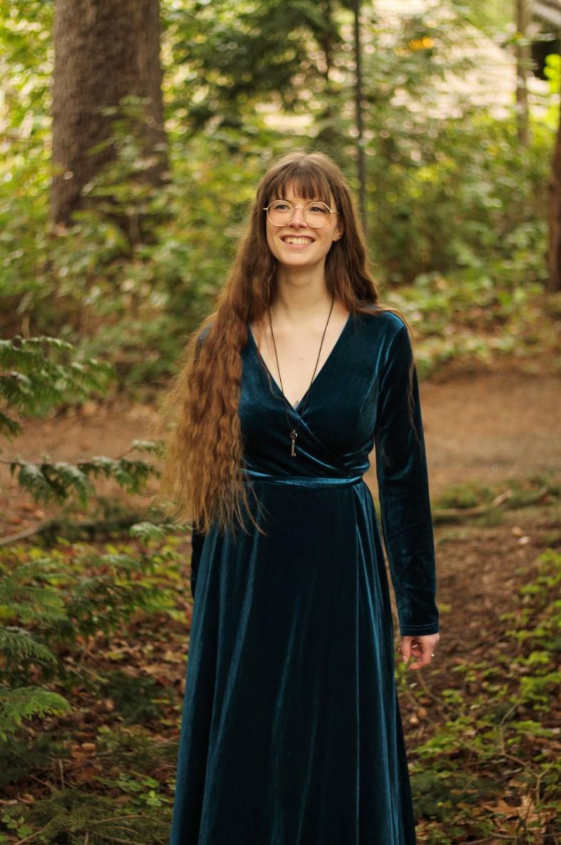 Caitriona Cassel standing in a forest with a big smile, wearing a long velvet green dress, waist length hair and round-lens glasses