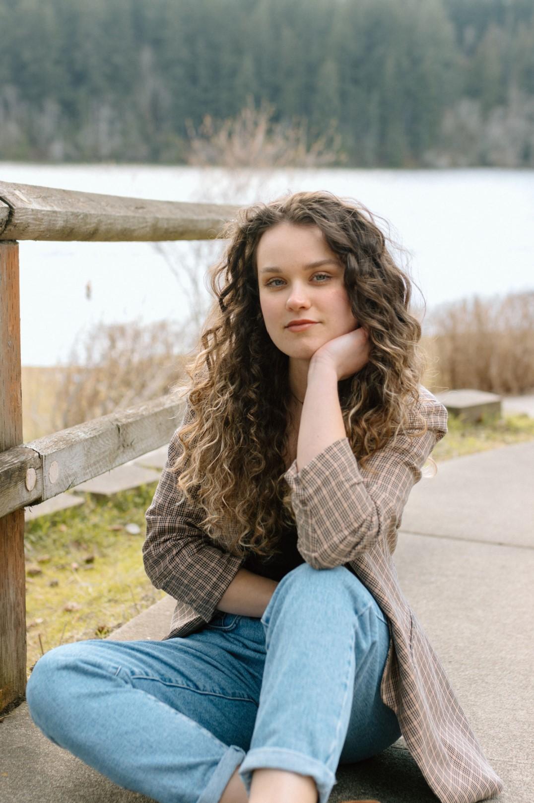 A person with long, curly hair resting their head on their hand, elbow on their knee, sitting on cement next to a fence by a lake.