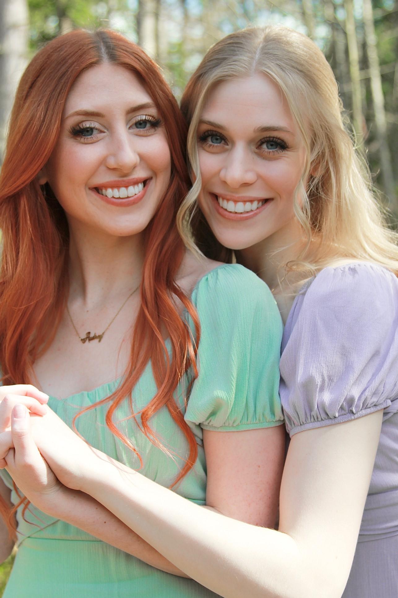 two girls with long hair: a red-head and a blond, embraced back to front in a friendly manner, and smiling