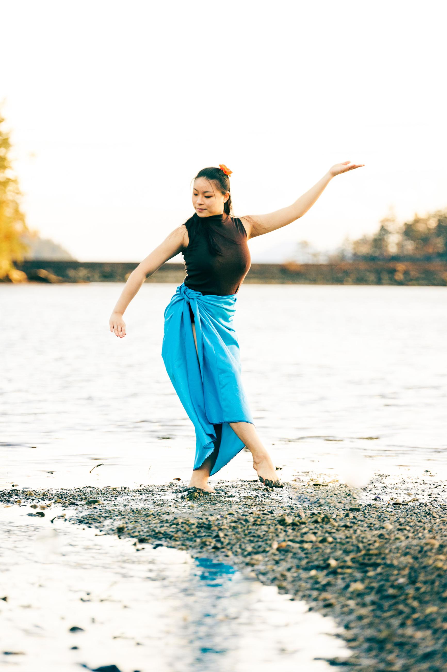 A person wearing a wrap-around skirt in a casual dance position with peaceful expression on a river sandbar