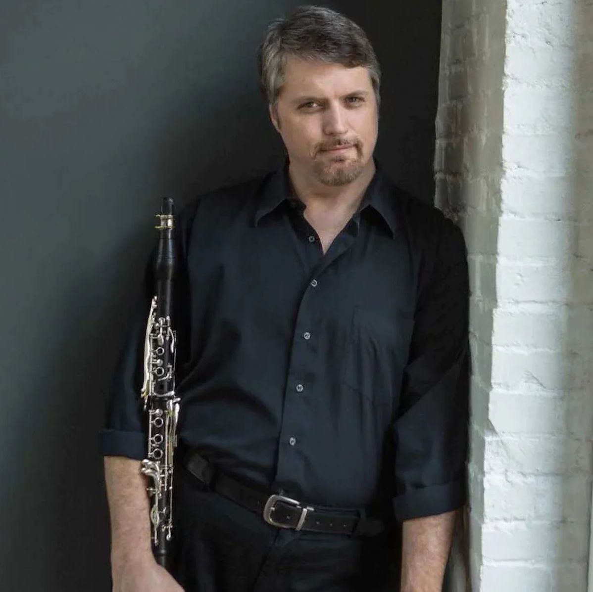 Sean Osborn leaning his shoulder against a brick wall, holding a clarinet in the opposite arm, sporting short graying hair and a trim goatee