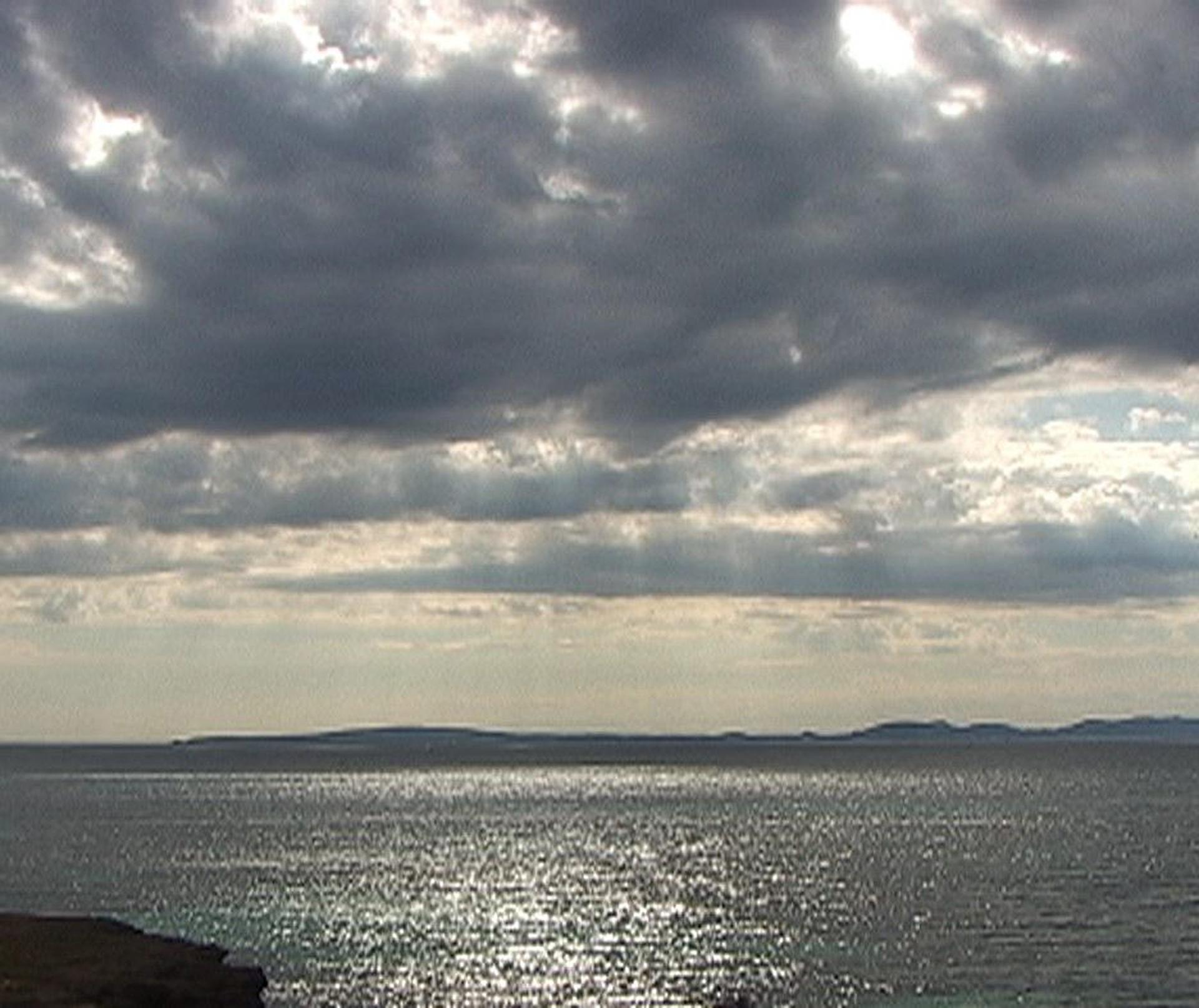 Sunrays shine down from gray clouds onto a bumpy ocean. Silhouetted islands appear on the horizon.