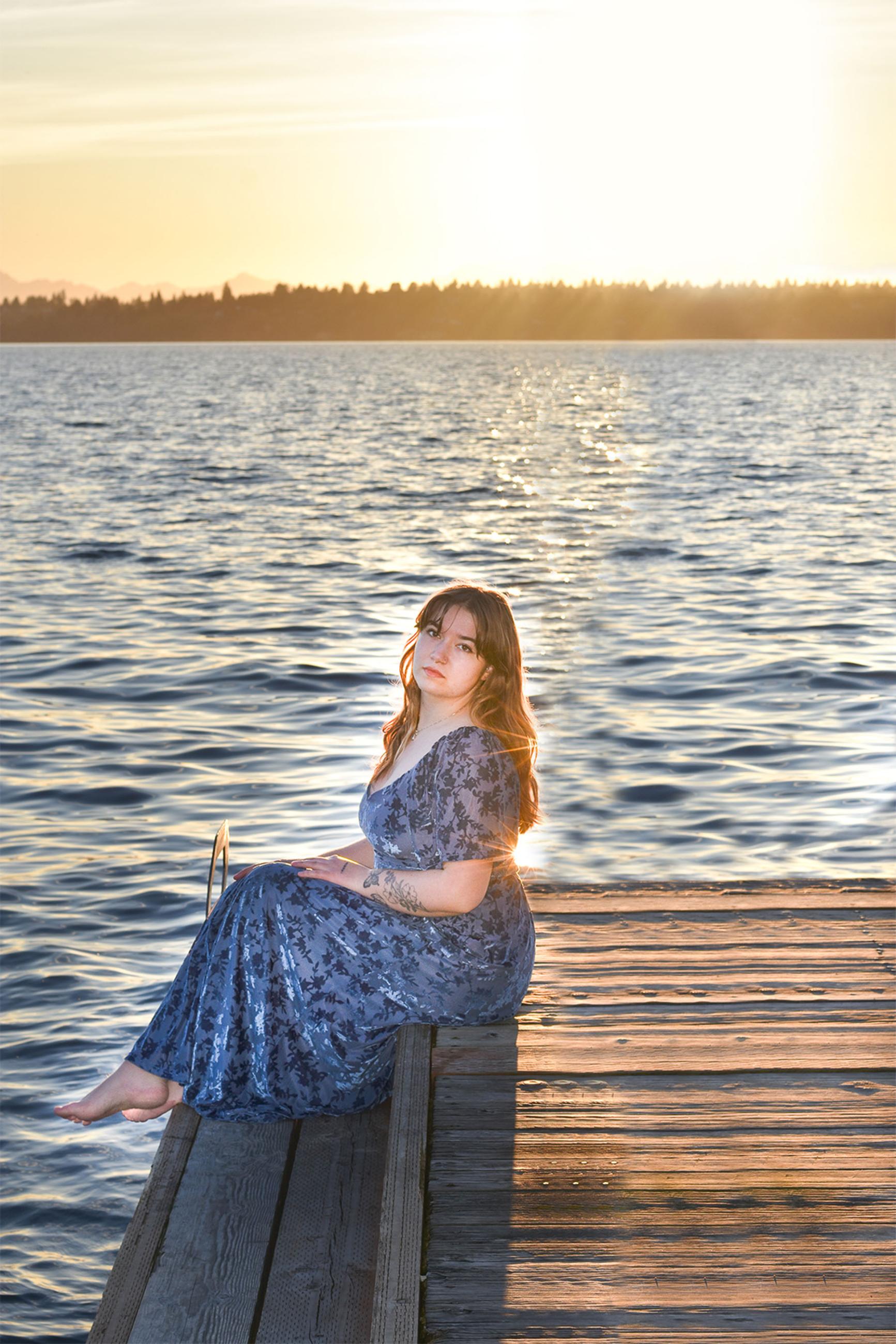 a person in a dress sits on a dock extending into the water with the sun setting in the background