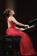 Rachel Cheung plays piano with gusto. She wears a long red sleeveless dress.