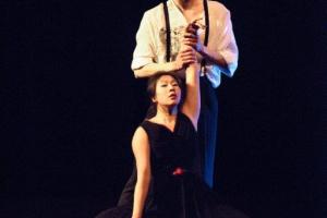 male dancer in suspenders and white shirt holds the arm of a female dancer in a dark dress, kneeling beneath him