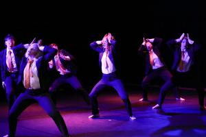 six performers wearing suits in saturated light. Some wear blindfolds