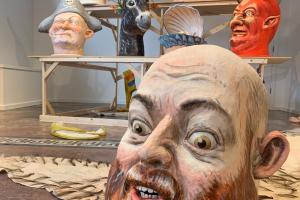 A giant head of Ryan Kelly. Behind are carnivalesque caricatures of animals, demons, witches