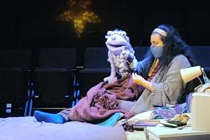 A person wearing a mask sits on a bed with a puppet. Behind them are empty theatre seats and a faintly lit star.