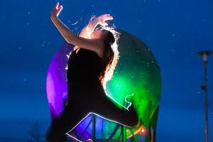 A backlit dancer leaps high and arches back with arms and legs curled. In the background is the colorfully-lit acid ball from the Bellingham waterfront park, under a starry night sky.