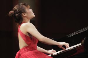 Rachel Cheung plays piano with gusto. She wears a long red sleeveless dress.