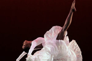 a sabel background frames a black female dancer in a form fitting bodice flinging her head and arms back so far they reach toward the floor. Her leg extends from the floating pinwheel of voluminous white fabric comprising her skirt.