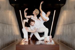 three dancers in white strike low, angular poses in a small space of concrete and wood with modern lighting and details