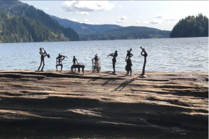 a sextet of small musician figurines sit on a driftwood log near a lake with mountains behind