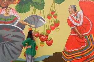 a woman stands in front of a brightly painted mural of Mexican folk dancers