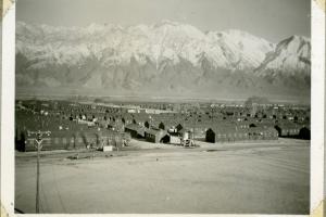 An aged black and white snapshot of ranks of barracks buildings against steep, aired snowcapped mountains.