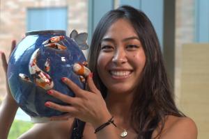 a person smiling broadly holds a blue ceramic olla near their head. The jug is blue and decorated with poi.