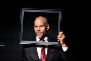 a man in a suit looks at us through an empty picture frame