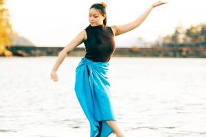 A person wearing a wrap-around skirt in a casual dance position with peaceful expression on a river sandbar